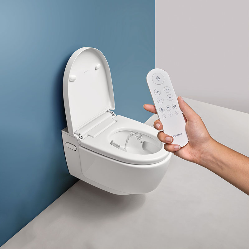 Inodoro bidé Geberit AquaClean: la mano de una mujer controla desde un mando a distancia el Inodoro bidé Geberit AquaClean suspendido en una pared en color azul cielo.