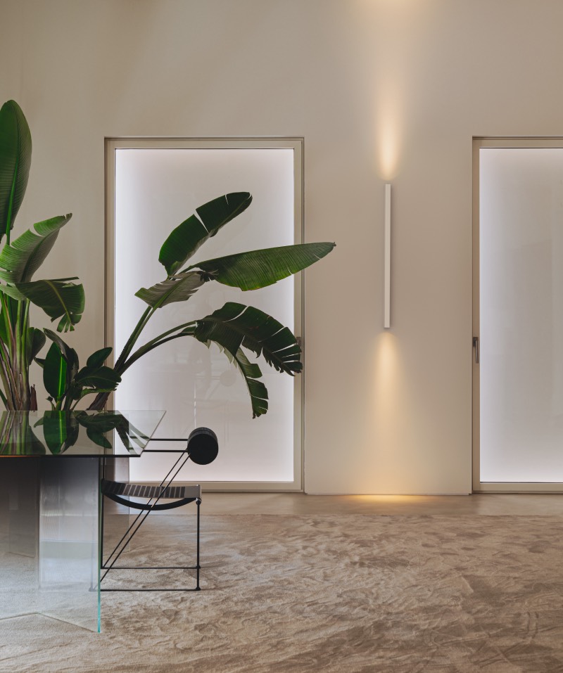 Offset: zona acogedora en colores tierra con una planta grande y una luminaria de pared de la colección Offset para alumbrar el espacio de manera indirecta.