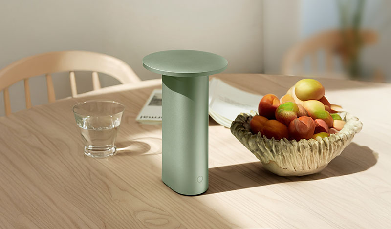 La Solae Lamp de Cecilie Manz para Fritz Hansen se muestra en tono verde sobre una mesa de madera junto a un cuenco de frutas y un vaso de agua