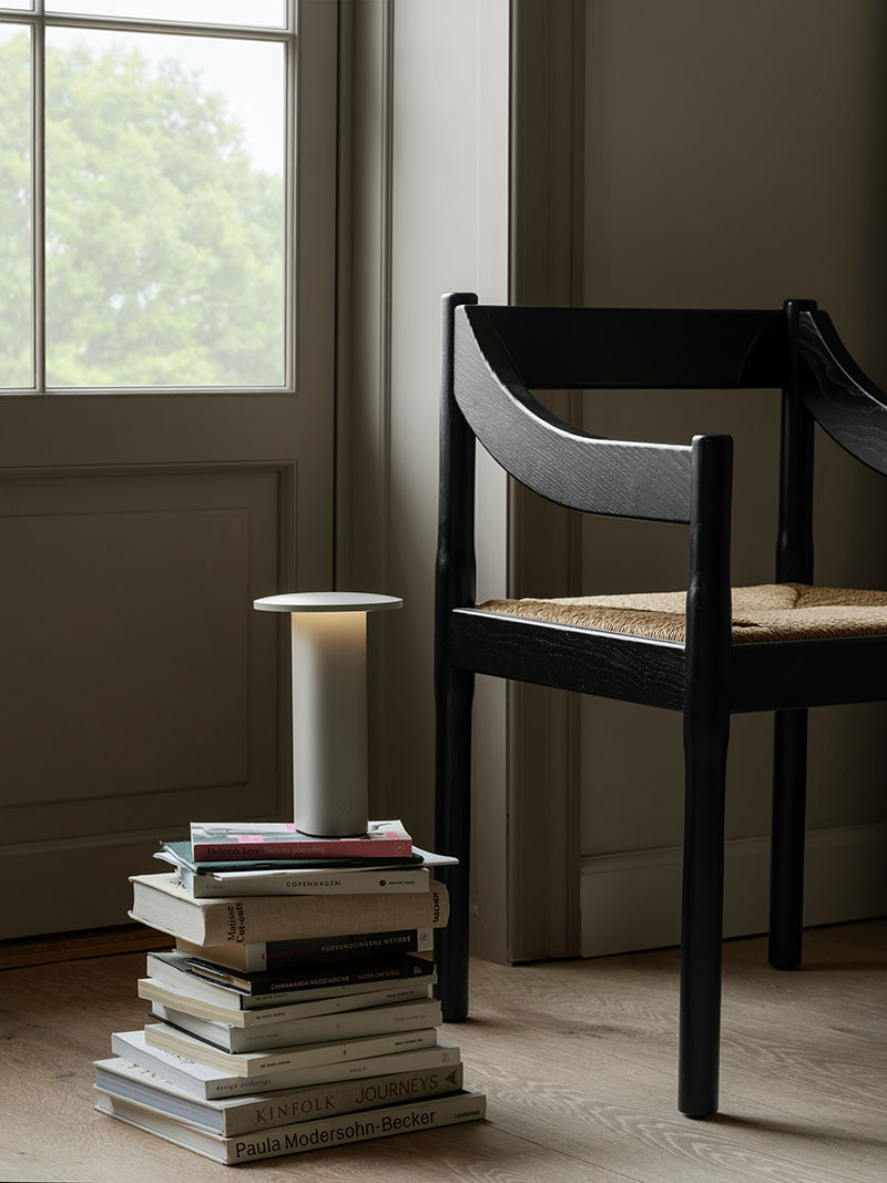 La Solae Lamp de Cecilie Manz para Fritz Hansen aparece encendida sobre una pila de libros al lado de una silla negra