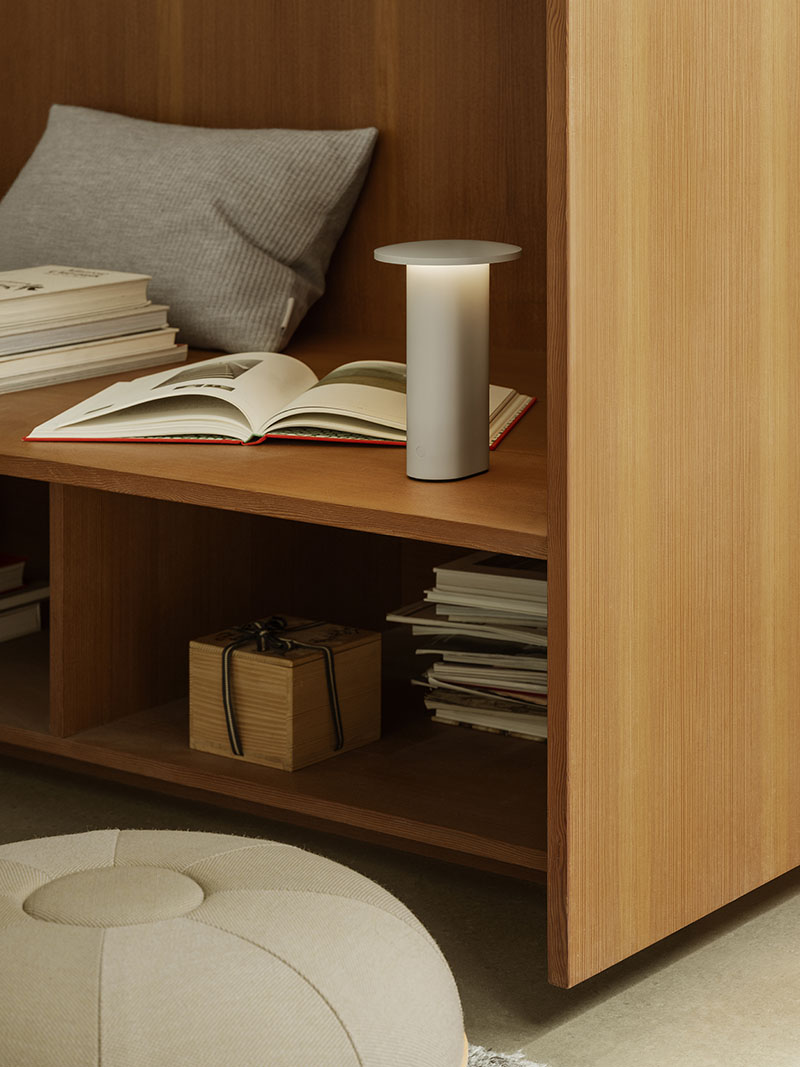 La Solae Lamp de Cecilie Manz para Fritz Hansen está colocada en una repisa de madera junto a un libro abierto y una almohada gris