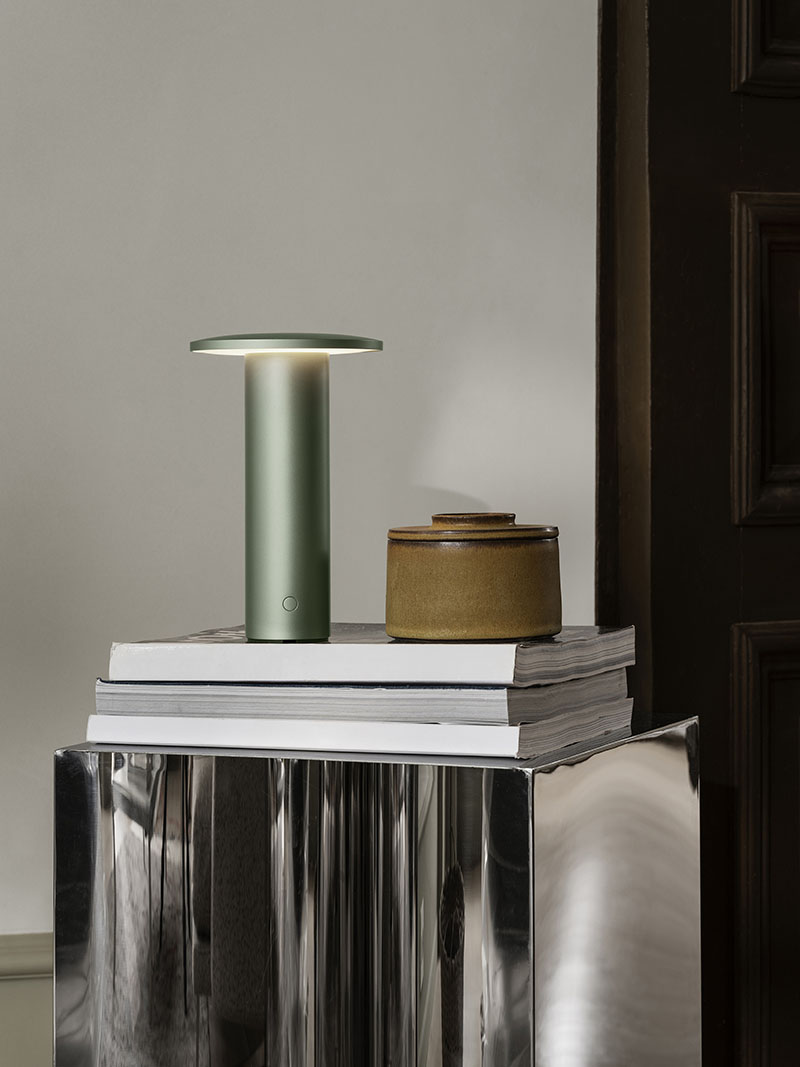 La Solae Lamp de Cecilie Manz para Fritz Hansen se observa en color verde sobre una pila de libros junto a un recipiente cerámico
