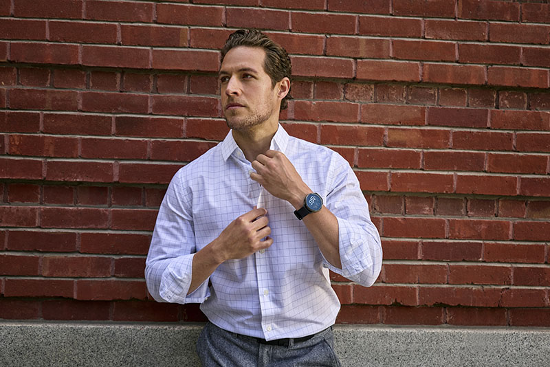 Venu 4: un hombre con camisa blanca de cuadros se coloca el cuello con sus manos en un espacio exterior con una pared de ladrillos detrás. En su muñeca izquierda luce un reloj inteligente Garmin Venu 4.