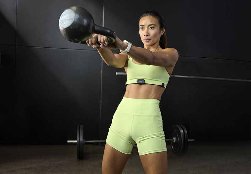 Venu 4: una mujer con una coleta y un conjunto deportivo en color verde lima realiza un entrenamiento de fuerza en un espacio oscuro y en su muñeca izquierda lleva un reloj inteligente Garmin Venu 4 con correa blanca.