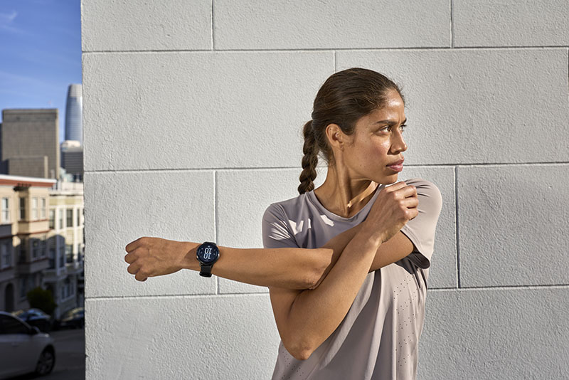 Venu 4: una mujer con una coleta trenzada y una camiseta deportiva en color gris realiza un estiramiento de brazo en un espacio exterior y en su muñeca izquierda lleva un reloj inteligente Garmin Venu 4 con correa negra.