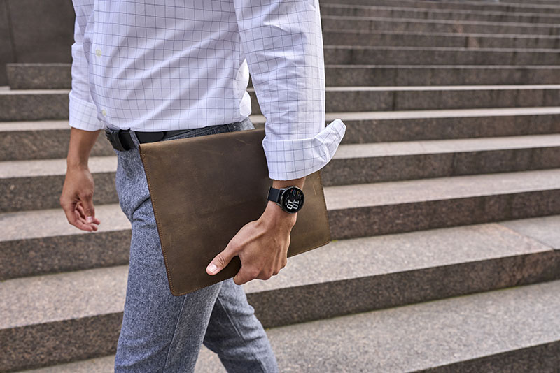 Venu 4: un hombre con camisa blanca de cuadros y pantalón camina por una zona exterior de escaleras con una carpeta en la mano con un reloj inteligente Garmin Venu 4.