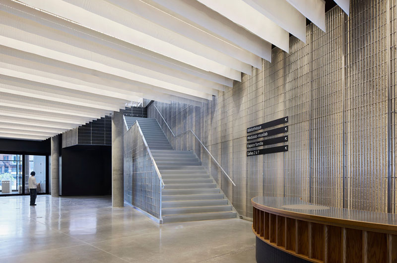 La escalera principal del Centre Sanaaq asciende junto a muros metálicos perforados bajo un techo de lamas blancas