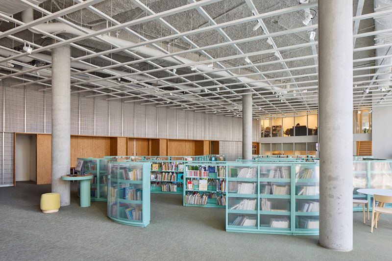 La zona infantil del Centre Sanaaq muestra estanterías bajas de color turquesa organizadas entre pilares de hormigón y muros metálicos