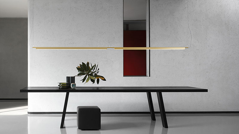 Compendium Suspension Luceplan: Espacio en tonos gris perla con una mesa negra grande que tiene una planta encima y un taburete debajo, sobre la que se encuentra la lámpara Compendium Suspension de Luceplan en color dorado.