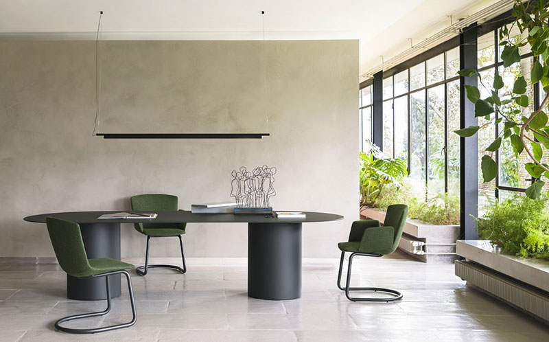 Compendium Suspension Luceplan: espacio luminoso con cristaleras en las que hay plantas, con paredes en tonos arena y con una mesa grande negra que tiene una figura decorativa y otros objetos de papelería encima, rodeada de tres sillas verdes y sobre la que se encuentra la lámpara Compendium Suspension de Luceplan en color negro.