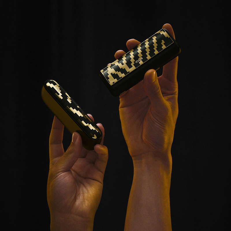 Dos manos elevan estuches de Iqos Iluma i x Seletti con el motivo geométrico de la colección