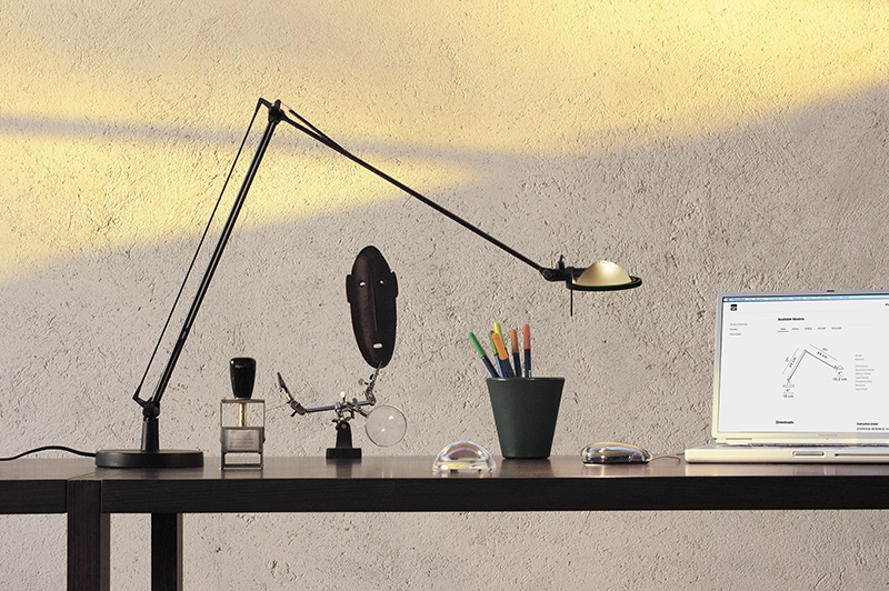 Luceplan: en una habitación iluminada por el sol se ve un escritorio negro, situado contra una pared en tono claro, con una lámpara de mesa con estructura negra Luceplan junto a un portátil blanco, un cubilete con rotuladores de colores y otros utensilios.
