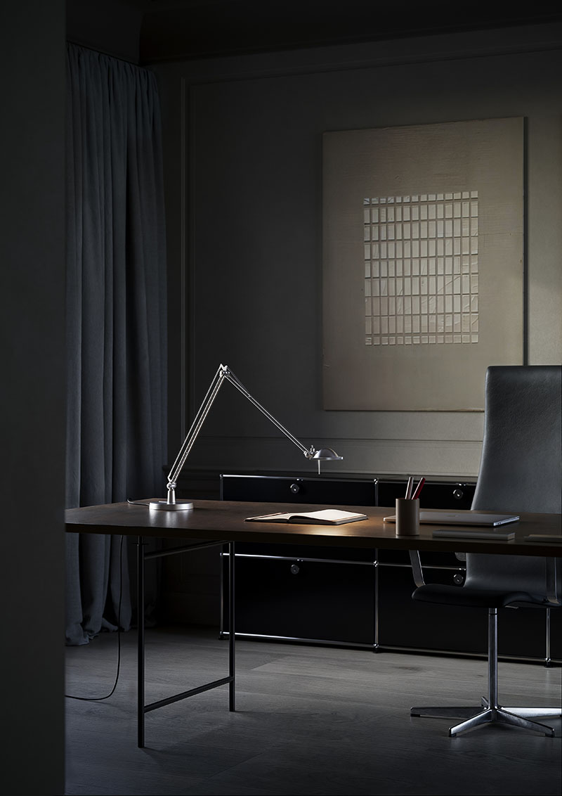 Luceplan: en una habitación oscura se ve un escritorio negro con una lámpara de mesa Luceplan encendida sobre un cuaderno y junto a un portátil negro, un cubilete con bolígrafos y una elegante silla negra detrás.