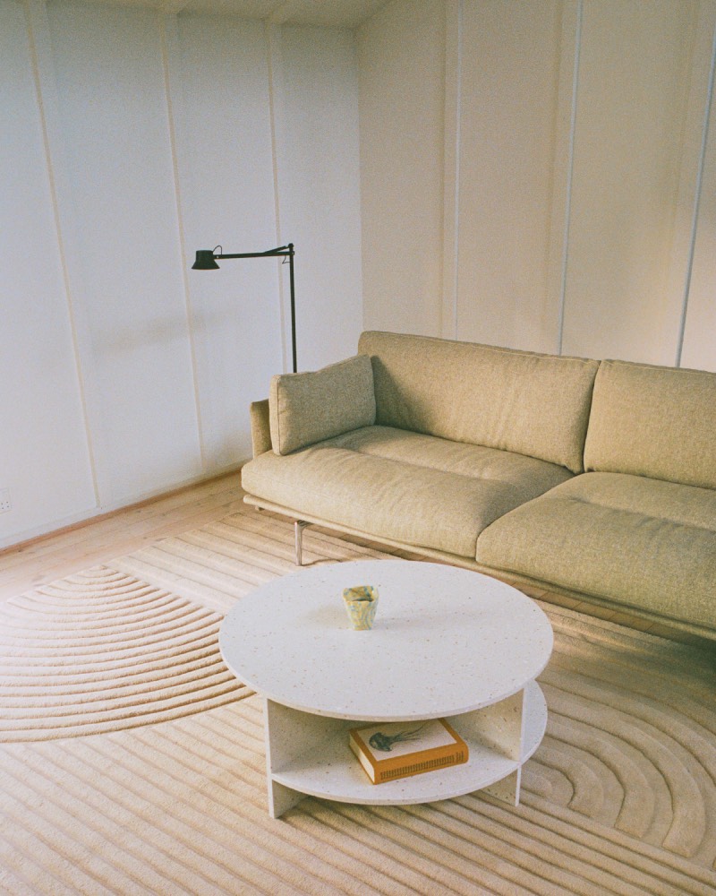 Muuto Halves MSDS Studio: salón en tonos claros con una lámpara de pie negra junto a un sofá en color tierra y una mesa de centro redonda sobre una alfombra con un dibujo lineal curvado en relieve.