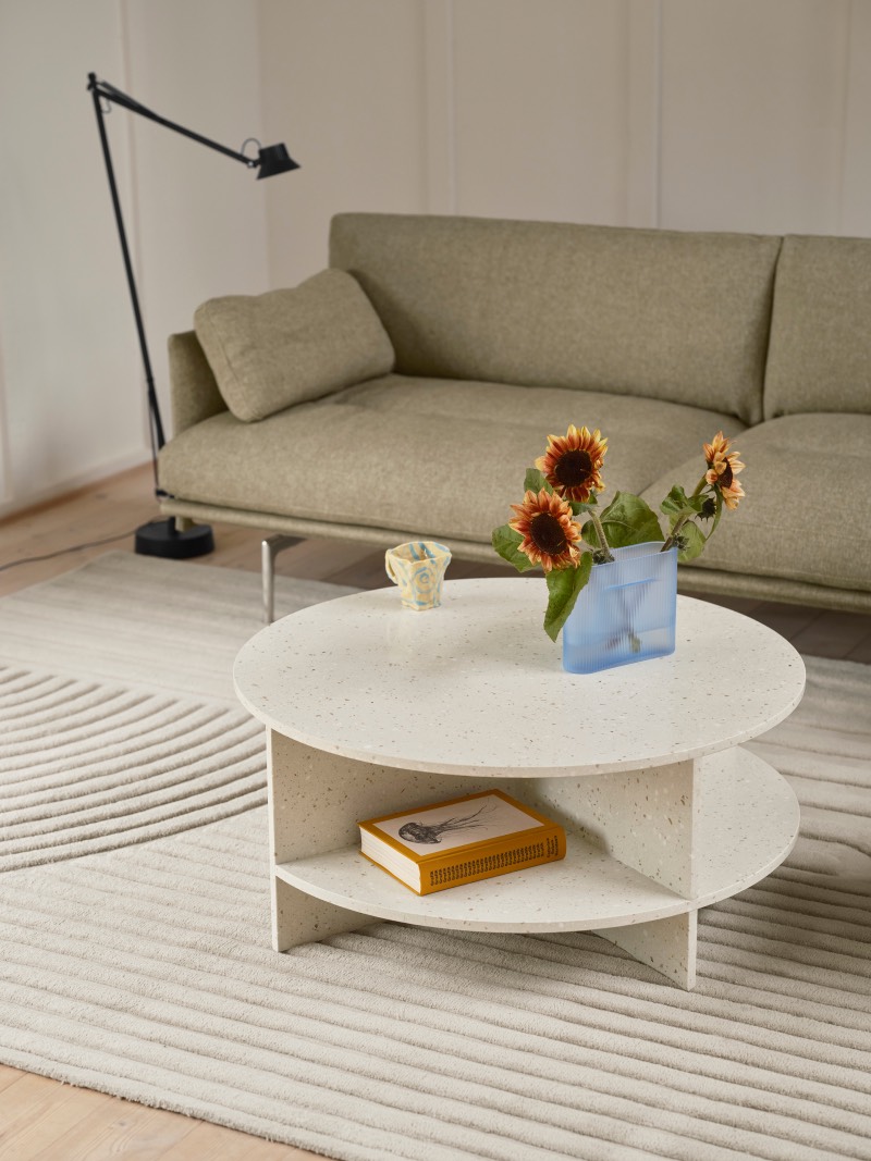 Muuto Halves MSDS Studio: salón en tonos claros con una lámpara de pie negra junto a un sofá en color tierra y una mesa de centro redonda, con unos girasoles encima y un libro en la parte baja, que reposa sobre una alfombra con un dibujo lineal curvado en relieve.