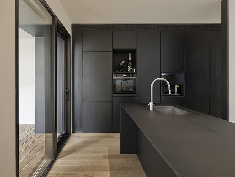 QB Atelier define en PDS House la cocina en isla con superficies oscuras y acceso acristalado a la terraza