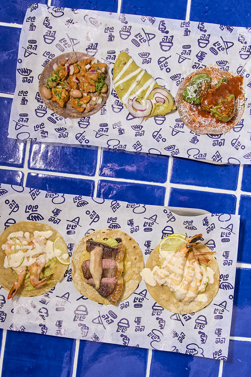 Lolo Taco Bar y Carne presentan una selección de tacos dispuestos sobre papel corporativo en una superficie de azulejo azul